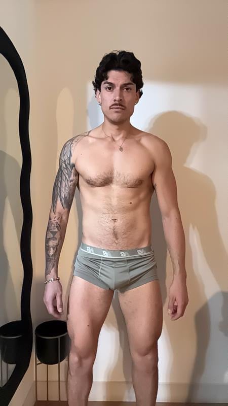 Shirtless man with dark hair and full sleeve tattoo on left arm standing in neutral pose against beige wall