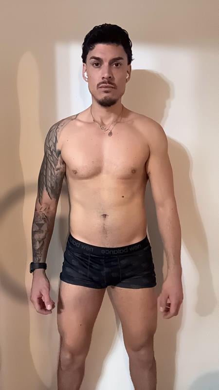 Shirtless man with dark hair, facial hair, and full-sleeve tattoo on left arm wearing black underwear against neutral wall