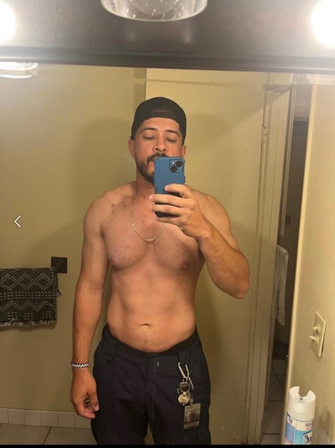 Shirtless man taking a mirror selfie with a blue phone, wearing a black cap and dark pants