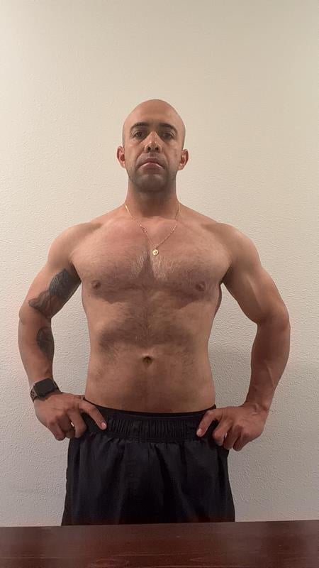 Shirtless man with muscular physique wearing black shorts, standing against a neutral beige wall in a front-facing pose