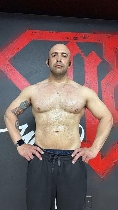Shirtless man posing with hands on hips against red and gray geometric wall art background, wearing black shorts and smartwatch